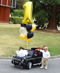 a little boy standing next to a black car with balloons in the shape of number one