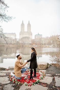 a man kneeling down next to a woman on top of a stone walkway near a lake