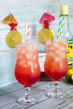 two glasses filled with drinks sitting on top of a wooden table next to bottles and limes