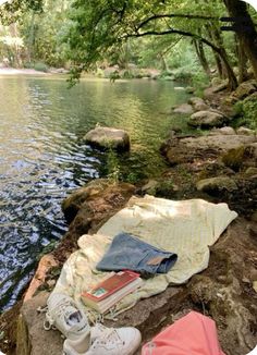 there is a blanket, book and shoes on the rocks by the riverbank with trees in the background