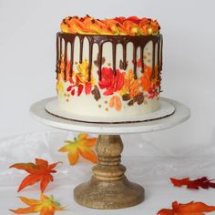 a white cake with chocolate frosting and fall leaves on it sitting on a pedestal