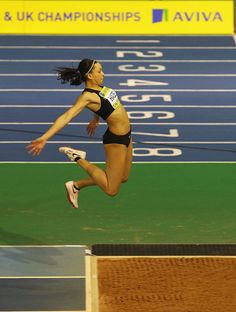 a woman jumping in the air on top of a blue and green track with an olympic sign behind her