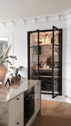 a kitchen with white walls and wooden flooring has an open glass door to the pantry