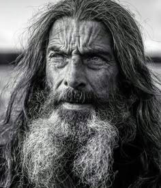 an old man with long hair and beard looks into the camera while standing in front of water