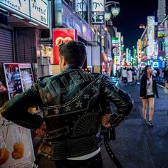 a man standing on the side of a street next to tall buildings at night with people walking by