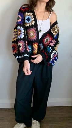 a woman standing in front of a white wall wearing black pants and a colorful crocheted sweater