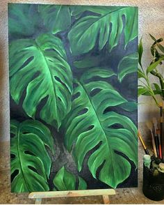 a painting with green leaves on it next to a potted plant and pencils