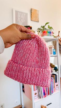 a hand holding a pink knitted hat in front of a bookshelf and shelves