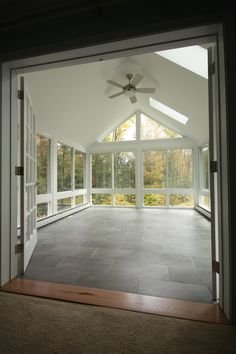 an empty room with large windows and a ceiling fan in the center, looking out onto trees