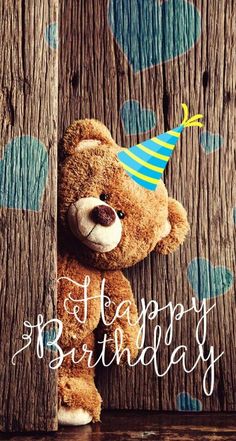 a brown teddy bear wearing a party hat peeking out from behind a wooden wall with the words happy birthday written on it