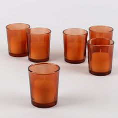 six orange glass tumblers sitting next to each other on a white surface with one candle in the middle