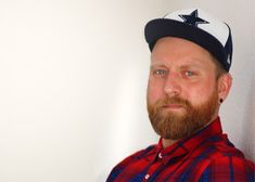 a man with a beard wearing a red and blue plaid shirt is standing in front of a white wall