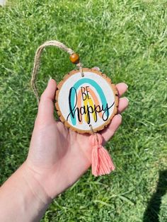 a hand holding a wooden ornament with the words be happy on it and a tassel