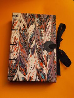 a notebook with an orange background and black ribbon on the cover is decorated with colorful feathers