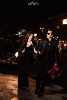 a man and woman dressed in black walking down the street at night with their arms around each other