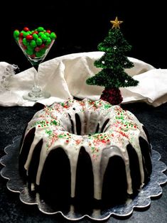 a bundt cake with white icing and sprinkles on it, next to a small christmas tree