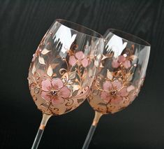 two wine glasses with pink flowers on them sitting side by side in front of a black background