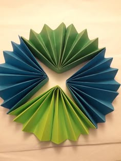 three folded green and blue paper fans on a white sheeted surface, with one folded in the shape of a flower