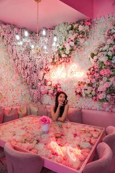 a woman sitting at a pink table with flowers on the wall behind her and chandelier above it