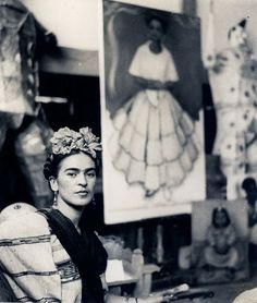 black and white photograph of a woman sitting in front of an easel with paintings on the wall behind her