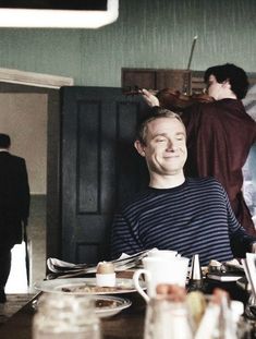 a man sitting at a table with food in front of him and two other people behind him