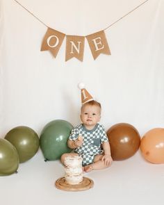 a baby sitting on the floor in front of balloons and a birthday cake with one sign above it