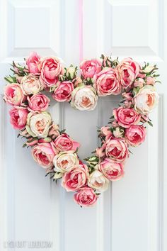 a heart shaped wreath with pink and white flowers hanging on the front door, decorated with greenery