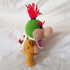 a crocheted toy is sitting on a white sheet and it looks like he's wearing a green hat with red hair