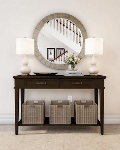 a table with baskets under a mirror and two lamps on top of it in front of a staircase