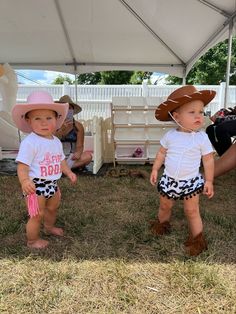 two toddlers wearing cowboy hats and shorts under a tent at an outdoor event with people sitting in the background