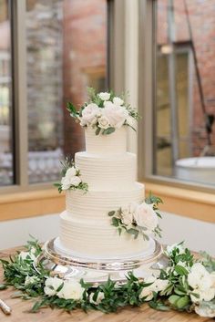 a wedding cake with white flowers and greenery sits on a table in front of a window