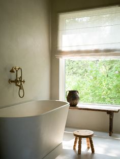 a white bath tub sitting under a window next to a wooden table and stool with a vase on it