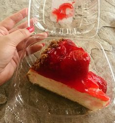 a hand holding a piece of cheesecake with strawberry sauce on it and ice cubes in the background