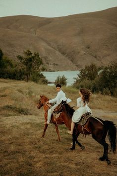 two people riding on the backs of horses in a field near water and hills behind them