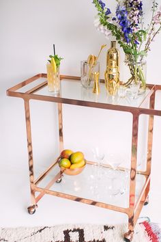 a glass and metal bar cart with drinks on it in front of a floral arrangement