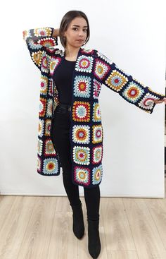 a woman standing in front of a white wall wearing a colorful crocheted cardigan