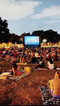 people are sitting on blankets in the grass watching a movie at an outdoor event with a large screen behind them