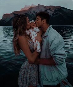 a man and woman kissing while holding a baby in front of the water with mountains behind them