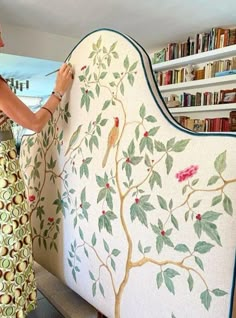 a woman is painting a wall with flowers and birds on it in front of a bookshelf