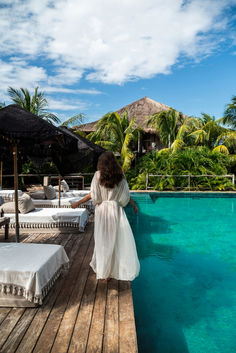 a woman in a white dress standing on a wooden dock next to a pool