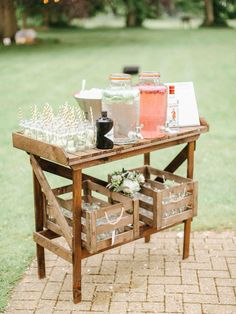 a wooden table topped with bottles and glasses filled with liquid on top of a field