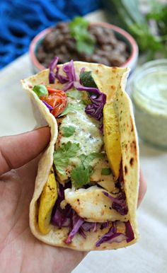 a hand holding a tortilla filled with meat and veggies next to a bowl of beans