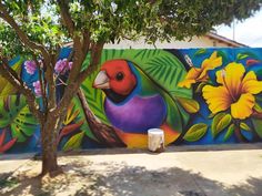 a colorful bird painted on the side of a building next to a tree and flowers