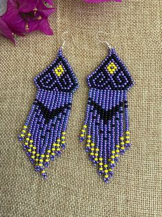 two purple and yellow beaded earrings on top of a table next to pink flowers