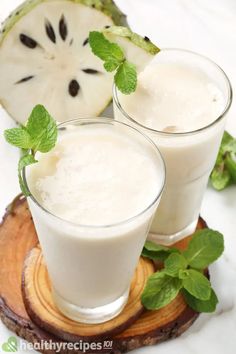 two glasses filled with white liquid sitting on top of a wooden coaster next to sliced kiwi