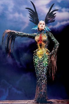 a woman dressed in an elaborate costume with feathers on her head and hands outstretched, standing against a cloudy background