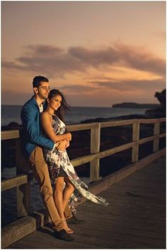 a man and woman are standing on a pier by the ocean at sunset with their arms around each other