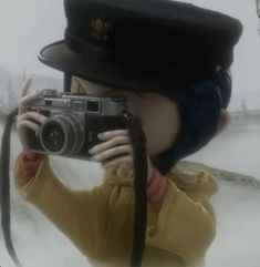 a doll is holding a camera in front of her face while wearing a hat and coat