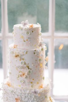 a three tiered white wedding cake with flowers on the top and bottom, sitting in front of a window