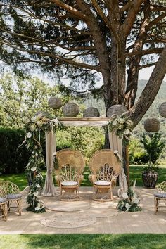 an outdoor wedding setup with wicker chairs and greenery on the ground under a large tree